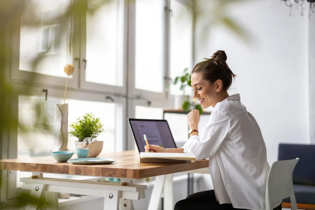 Smilende kvinne skriver i notatbok ved skrivebord med laptop