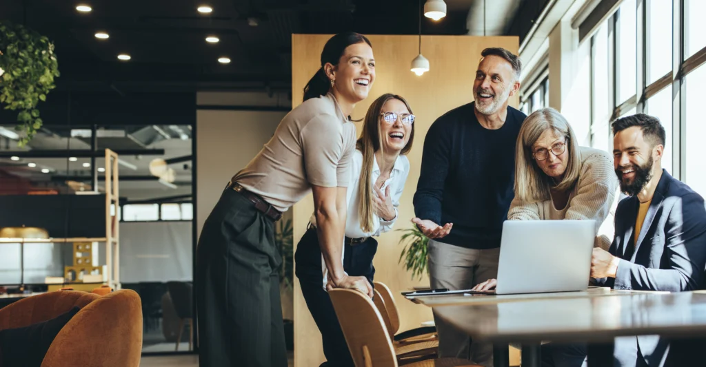 Smiling coworkers gathered around laptop in modern office collaboration space